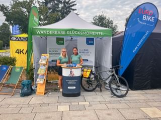Two staff members in turquoise shirts at an Eurobike promotional tent with banners and a bicycle. Green banner reads 'Wanderbare Glücksmomente'.