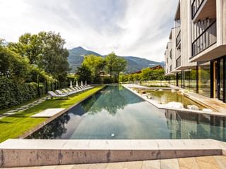 Moderner Außenpool im Hotel Sand mit Liegestühlen auf grüner Wiese, umgeben von Bergen und Bäumen in Südtirol.