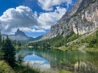 Lago Federa reflecting dramatic Dolomite peaks and clouds. Pine forests surround the green alpine lake with steep rocky cliffs on the right.