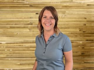 Portrait of Tamara Gerner smiling warmly, wearing a gray polo shirt, standing in front of a natural wooden wall.