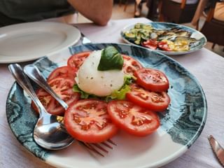 Caprese salad with sliced tomatoes, mozzarella, and basil on a blue plate. Grilled vegetables visible on a second plate in the background.