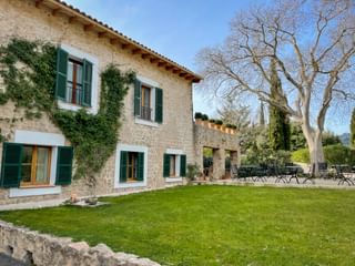 Steinfassade des Hotels Son Palou Orient mit grünen Fensterläden, efeubedeckten Wänden, Terrasse mit Stühlen und gepflegtem Rasen unter blauem Himmel.
