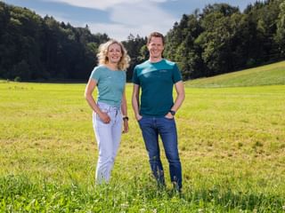 Two people Verena Sonnenberg und Thomas Schmid standing in a green meadow with forest in the background. Both wear blue outdoor shirts and smile at the camera.