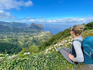 Female hiker with blue backpack sitting on hillside overlooking Madeira's north coast with dramatic cliffs, ocean, and white flowers in foreground.