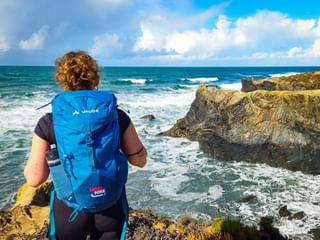 Wanderin mit blauem Rucksack betrachtet felsige Küste an der Praia das Furnas an der Rota Vicentina. Wellen brechen an goldenen Felsen unter blauem Himmel.