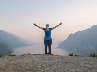 Woman with raised arms standing on rocky summit at Monte Brione overlooking Lake Garda at sunset, mountains visible on both sides.
