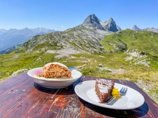 Two dessert slices on white plates at a mountain hut table, with green alpine meadows and rocky peaks of the Allgäu-Lechtal Alps behind.