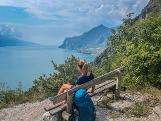 Wanderin mit Pferdeschwanz sitzt auf Holzbank mit blauem Rucksack und blickt auf Gardasee und Berge. Stadt am fernen Ufer sichtbar.