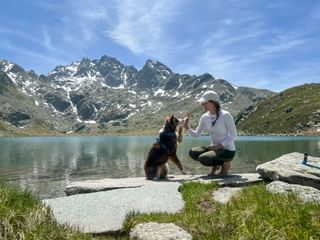 Woman in white shirt and cap crouching beside a dog on rocks at Rusetta Lake, with snow-capped mountains in the background on Via Spluga.