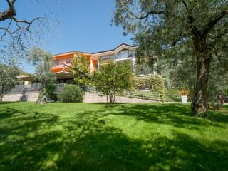 Hotel Catullo in Malcesine mit orange-weißer Fassade, umgeben von grüner Rasenfläche, Olivenbäumen und mediterraner Vegetation unter blauem Himmel.