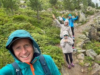 Smiling man taking selfie with family on rocky mountain trail in rain. Three people behind wear rain jackets, surrounded by green vegetation.