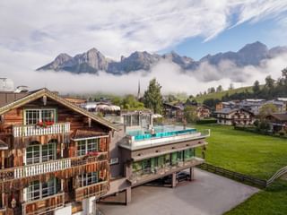 Hotel Sepp in Maria Alm with traditional wooden chalet and modern building with rooftop pool, surrounded by green meadows and Alpine peaks.