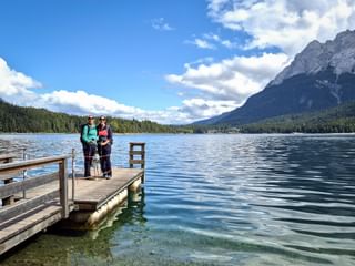 Two hikers standing on a wooden pier at Lake Eibsee with clear turquoise water, forested shores, and Zugspitze mountain in the background.