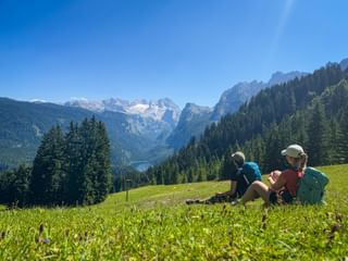 Two hikers with backpacks sitting on a green meadow in Gosau, viewing the Dachstein glacier and mountain peaks under a clear blue sky.
