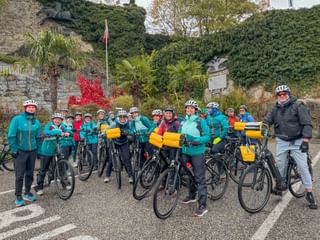 Große Gruppe von Radfahrern in türkisfarbenen Jacken mit Helmen und Fahrrädern mit gelben Taschen vor Steinmauer mit Palmen.