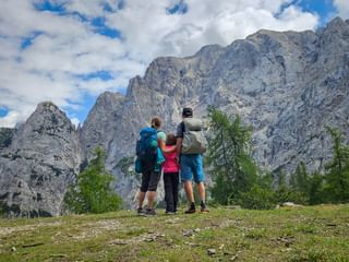 Family of three with backpacks viewing dramatic rocky peaks of the Julian Alps. Green meadow and trees in foreground, cloudy blue sky above.