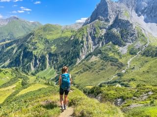 Hiker with blue backpack walking on mountain trail overlooking green valleys and rocky peaks in the Allgäu and Lechtal Alps.