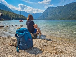 Hiker with blue backpack sitting on rocks at Lake Bohinj shore. Turquoise water, sailboat, and Julian Alps mountains under blue sky visible.