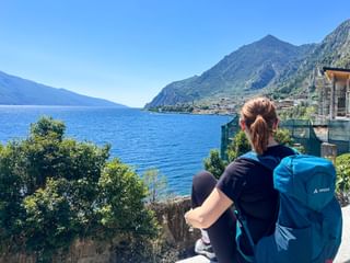 Hiker with blue backpack viewing Lake Garda and Limone sul Garda, with mountains and blue water visible under clear sky.