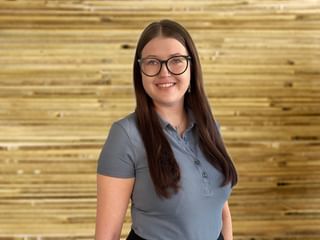Portrait von Magdalena Ottinger mit Brille und langen braunen Haaren, trägt ein graues Poloshirt und lächelt vor einer Holzwand.