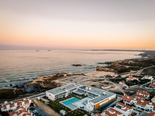 Luftaufnahme des Praia Hotel & Spa Porto Covo bei Sonnenuntergang mit weißem modernen Gebäude und Pool an felsiger Küste.