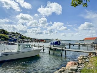 White motorboats moored at wooden piers in Särö harbor. Rocky shoreline in foreground, red-roofed building visible, blue sky with white clouds.