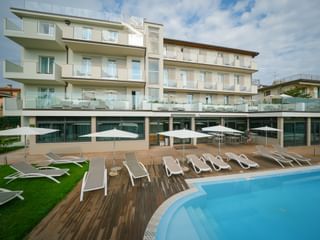 Modernes Hotel Palazzo del Garda Gebäude mit Glasbalkonen und Poolbereich mit weißen Sonnenliegen auf Holzterrasse.