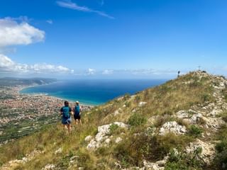 Two hikers walking on Monte Croce with panoramic view of the Ligurian coast and Mediterranean Sea. A summit cross is visible on the rocky peak.