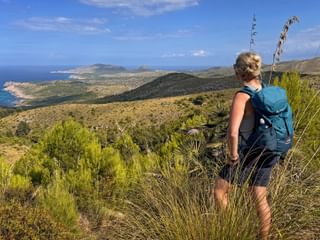 Wanderin mit blauem Rucksack auf Hügel mit Blick auf Mallorcas Ostküste mit türkisfarbenem Meer und bergiger Landschaft.