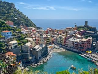 Aerial view of Vernazza in Cinque Terre with colorful houses along the harbor, a stone tower, and turquoise Mediterranean waters.