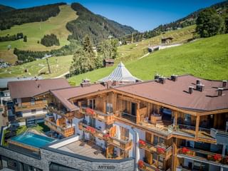 Aerial view of Hotel Astrid in Saalbach with wooden balconies and outdoor pool, surrounded by green Alpine meadows and ski slopes.
