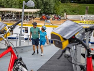 Two cyclists walking on boarding ramp of Danube river cruise ship in Melk. Bicycles visible in foreground, yellow ship railing with 'Rad & Schiff' text.