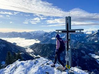 Hiker in purple jacket standing at summit cross on snowy peak, overlooking snow-covered Alpine valleys and mountain ranges under blue sky.