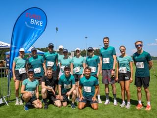 Gruppe von Läufern in türkisfarbenen Shirts mit Startnummern auf Rasen. Eurobike-Flagge links unter blauem Himmel sichtbar.