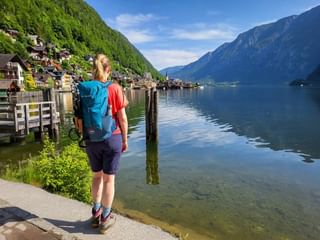 Female hiker with blue backpack viewing Hallstatt village across calm lake waters, surrounded by steep forested mountains under blue sky.