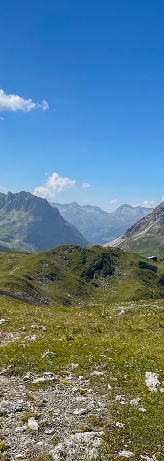 Steiniger Wanderweg durch grüne Almwiesen mit Berggipfeln in den Allgäuer Alpen unter blauem Himmel mit weißen Wolken.