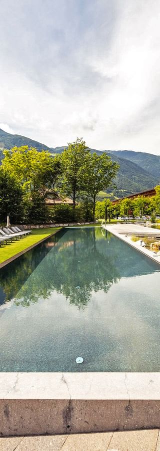 Moderner Außenpool im Hotel Sand mit Liegestühlen auf grüner Wiese, umgeben von Bergen und Bäumen in Südtirol.