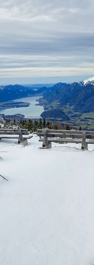 Wanderwegweiser im Schnee auf der Katrin mit gelben und blauen Richtungsschildern, Holzbänken und Panoramablick auf See und Berge.