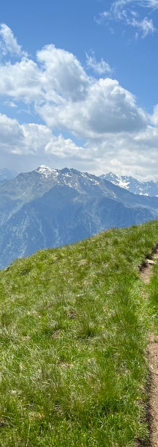 Wanderer in roter Jacke auf grasigem Bergweg mit Blick auf das Passeiertal und schneebedeckte Alpengipfel unter blauem Himmel mit Wolken.