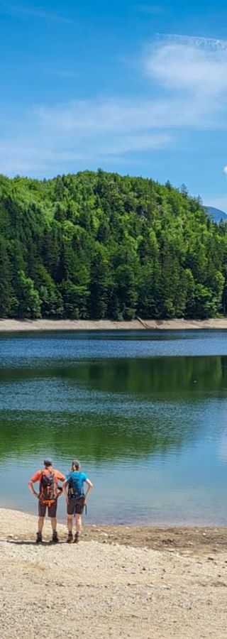 Two hikers with backpacks standing on sandy shore of Nussensee, surrounded by forested hills under blue sky with white clouds.