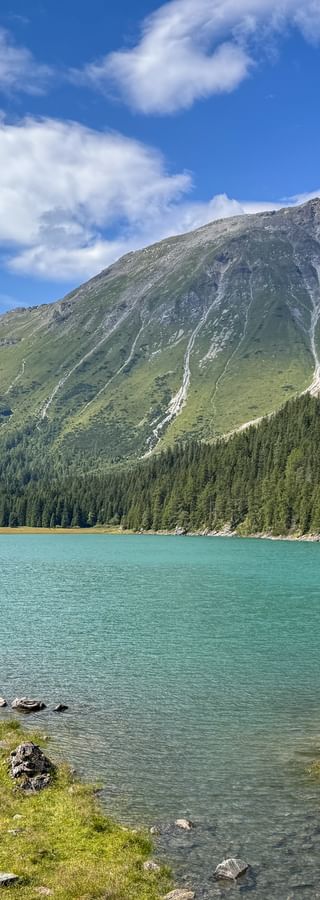 Türkisfarbener Obernberger See umgeben von Nadelwald mit dramatischen Berggipfeln im Hintergrund unter blauem Himmel mit weißen Wolken.
