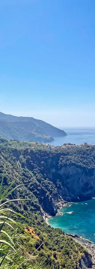 Panoramic view of Cinque Terre coastline with steep cliffs, terraced hillsides, and turquoise Mediterranean Sea under clear blue sky.