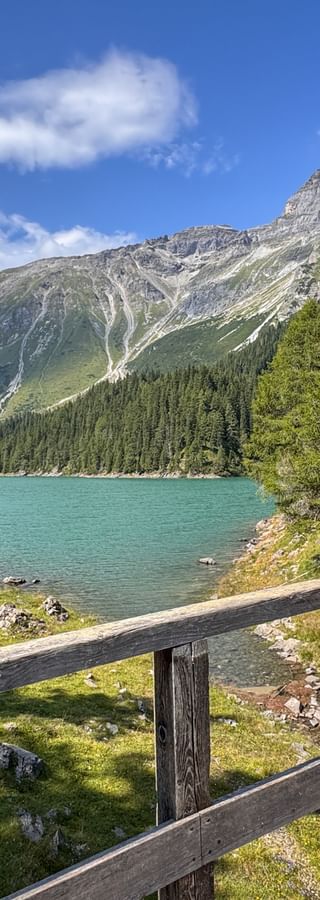 Wanderin mit rotem Rucksack und Wanderstöcken auf Holzbrücke mit Blick auf türkisfarbenen Obernberger See mit Bergen und Wald.