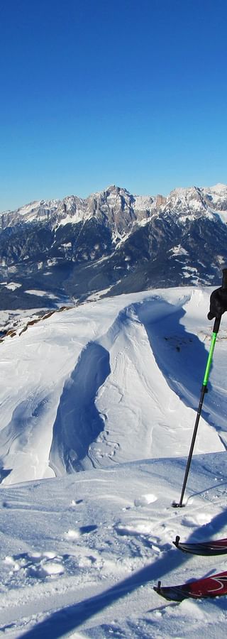 Skifahrer in blauer Jacke mit Stöcken auf verschneitem Grat am Hochkönig, mit schneebedeckten Berggipfeln und Tal unter blauem Himmel.