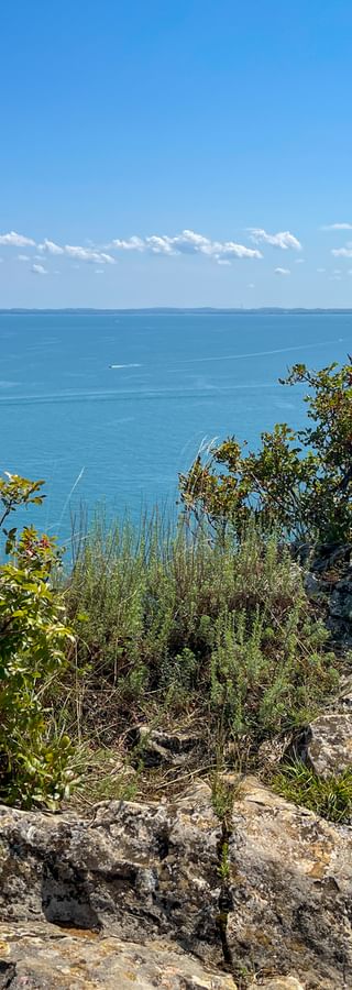 Felsige Klippe mit wilder Vegetation mit Blick auf das türkisfarbene Wasser des Gardasees unter blauem Himmel mit vereinzelten Wolken.