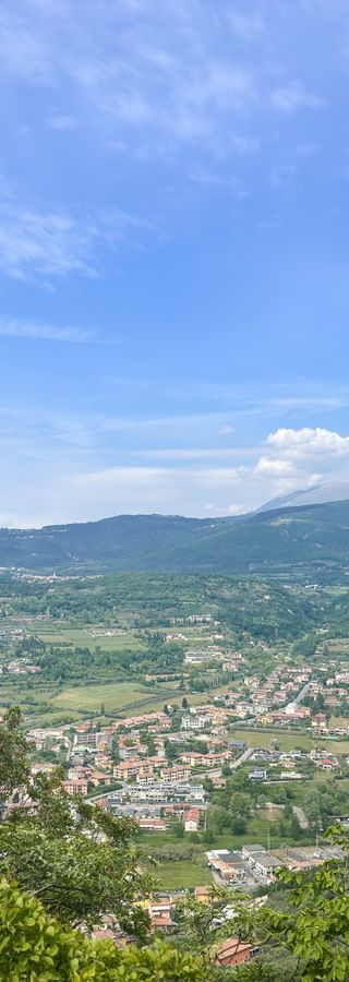 Panoramablick vom Hang über das Gardasee-Hinterland mit Ortschaften, grünen Feldern und Bergen unter blauem Himmel mit Wolken.