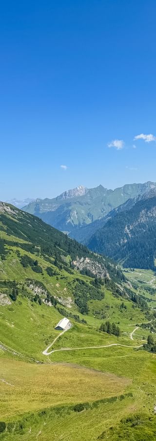 Panoramablick auf ein grünes Alpental mit gewundenen Wegen, umgeben von felsigen Berggipfeln unter blauem Himmel mit weißen Wolken.