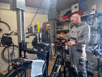 Two men in a bike workshop during Eurofun Touristik Training Days 2026. One holds an e-bike while the other demonstrates bike maintenance techniques.