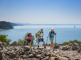 Three hikers with backpacks and walking poles standing on rocky terrain overlooking the Adriatic Sea near Trieste in the Julian Alps.