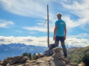 Hiker in blue shirt standing at rocky summit with wooden cross on Haideralm trail, overlooking mountain ranges and valleys under blue sky.